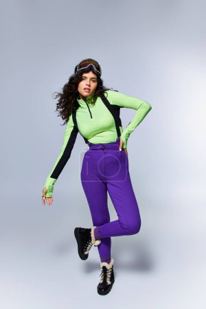winter sport, full length of sporty woman with curly hair posing in active wear on grey backdrop