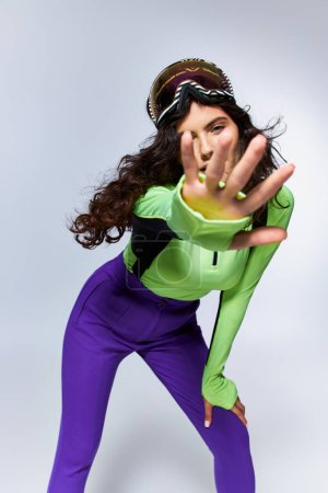 winter fashion, sporty brunette woman with curly hair posing in ski clothes with outstretched hand