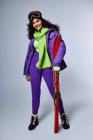 Foto de Deporte de invierno, mujer alegre con el pelo rizado posando en desgaste activo con chaqueta hinchable y esquís - Imagen libre de derechos