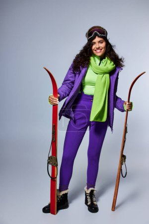 Foto de Deporte de invierno, mujer sonriente con el pelo rizado posando en desgaste activo con chaqueta hinchable y esquís - Imagen libre de derechos