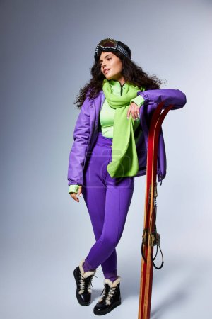Winteraktivität, charmante Frau mit lockigem Haar posiert in aktiver Kleidung mit Kugeljacke und Skiern