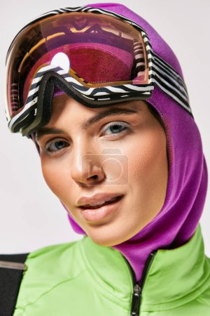 stylish woman in winter ski clothes with balaclava on head looking at camera on grey backdrop