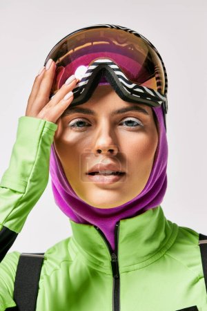 young woman in winter ski clothes with balaclava on head looking at camera on grey backdrop