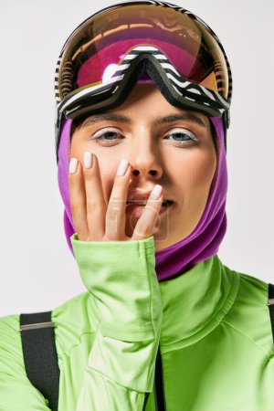 junge Frau in Winterskikleidung mit Sturmhaube auf dem Kopf posiert mit den Händen in Gesichtsnähe auf grau