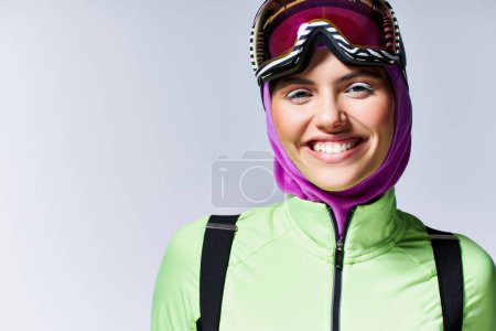 fröhliche Frau in winterlicher Aktivkleidung mit Sturmhaube auf dem Kopf und Skibrille vor grauem Hintergrund