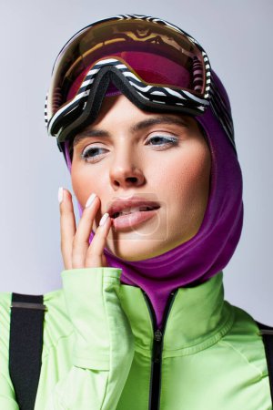 beautiful woman in winter ski clothes with balaclava on head posing with hand near face on grey
