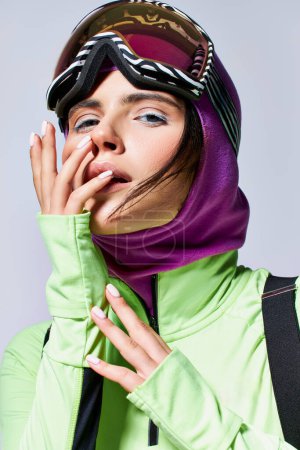 portrait of woman in ski clothes with balaclava posing on grey backdrop, model with blue eyes