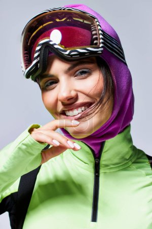 Porträt einer zufriedenen Frau in aktiver Kleidung mit Sturmhaube auf dem Kopf, die auf grauem Hintergrund lächelt
