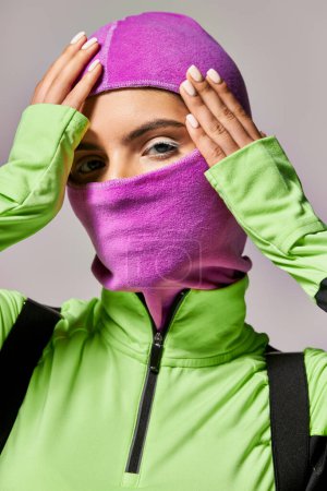 portrait of woman with blue eyes wearing purple ski mask and looking at camera on grey background