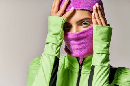 portrait of sportive woman with blue eyes wearing purple ski mask and looking at camera on grey