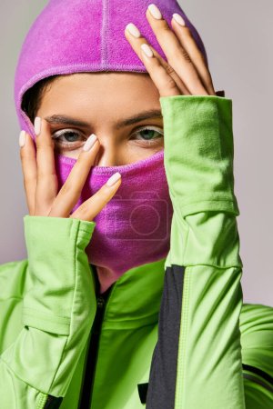 portrait of sportive woman with blue eyes wearing ski mask and looking at camera on grey backdrop