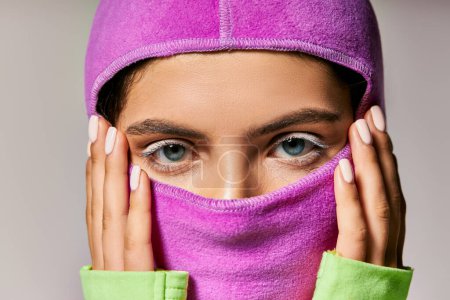 close up of young woman with blue eyes wearing ski mask and looking at camera on grey backdrop