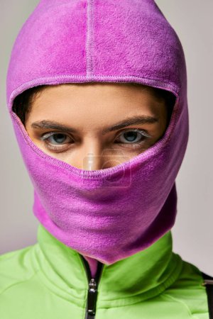 portrait of young woman with blue eyes in purple ski mask looking at camera on grey backdrop