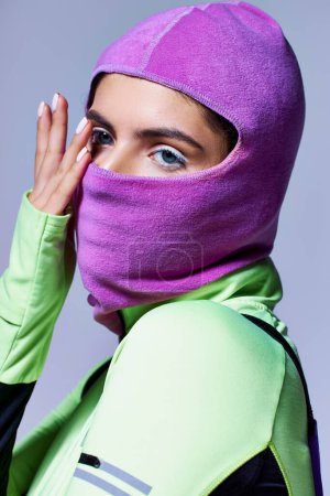 portrait of woman with blue eyes in warm ski mask posing with hand near face on grey backdrop