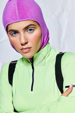 portrait of attractive woman with blue eyes posing in warm ski mask and active wear on grey backdrop