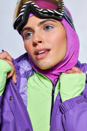 young woman with blue eyes posing in ski googles, balaclava and winter jacket on grey background