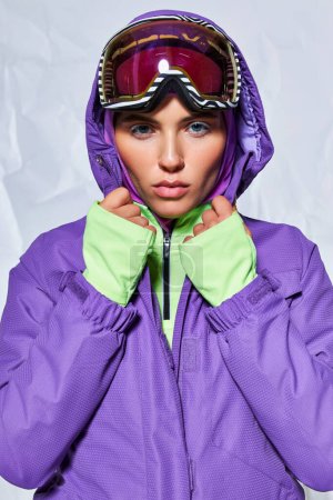 serious woman with blue eyes posing in ski googles, balaclava and winter jacket on grey background