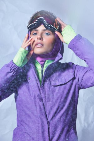 hermosa mujer en pasamontañas y chaqueta de invierno con googles de esquí sobre fondo gris, día nevado