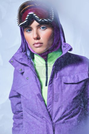 attractive woman in balaclava, purple winter jacket and ski googles posing on grey, snowing day
