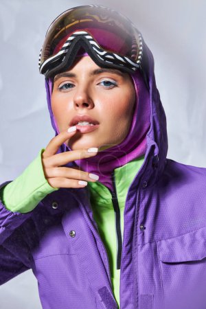 bonita mujer en pasamontañas, chaqueta de invierno púrpura y esquí googles posando con la mano cerca de la cara en gris