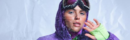 jolie femme en cagoule, veste d'hiver violette et googles de ski posant sur gris, bannière enneigée