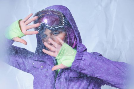 Téléchargez les photos : Jolie femme en cagoule, veste d'hiver violette et lunettes de ski posant sur gris, temps neigeux - en image libre de droit