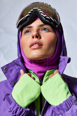 Kälte, verträumte Frau mit Sturmhaube und Skibrille posiert in lila Winterjacke auf grau