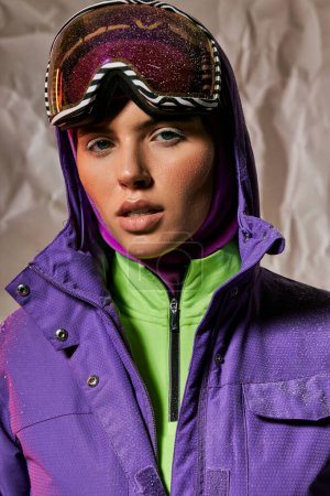aire fresco, hermosa mujer en pasamontañas y esquí googles posando en chaqueta de invierno púrpura sobre gris
