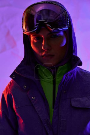 hermosa mujer en pasamontañas, esquí googles y chaqueta caliente posando sobre fondo púrpura, moda de invierno