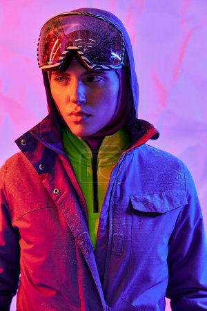 hermosa mujer en máscara de esquí y googles usando chaqueta caliente sobre fondo púrpura, moda de invierno