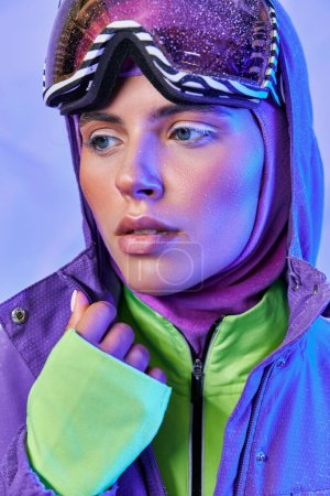 atractiva mujer en máscara de esquí, googles y chaqueta cálida mirando hacia otro lado en el telón de fondo púrpura, belleza de invierno