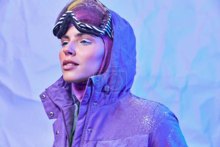 aire helado invierno, mujer joven en máscara de esquí, googles y chaqueta cálida mirando hacia otro lado en el fondo púrpura
