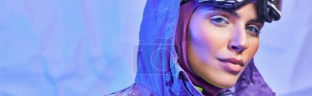 pancarta de invierno, mujer joven en máscara de esquí, googles y chaqueta caliente mirando a la cámara en el fondo azul