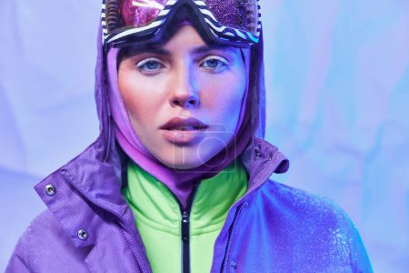 frostige Luft, junge Frau mit Skimaske, Brille und Winterjacke blickt vor blauem Hintergrund in die Kamera