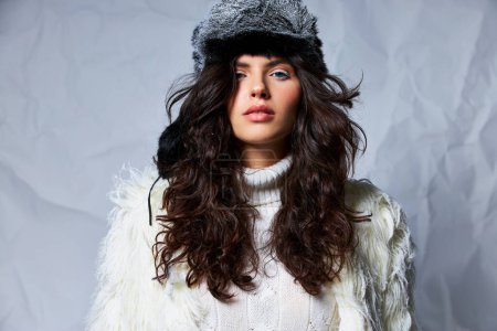 curly woman in faux fur hat and white sweater looking at camera on grey backdrop, winter beauty