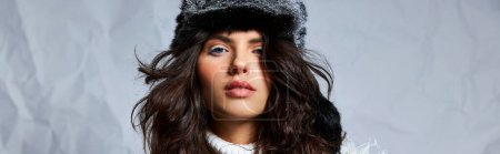 curly woman in faux fur hat and  sweater looking at camera on grey backdrop, winter beauty banner