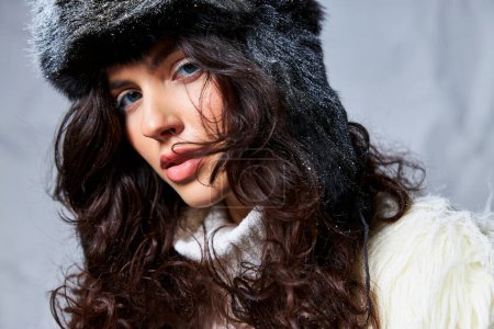 snow on furry hat, curly brunette woman in white sweater looking at camera on grey backdrop