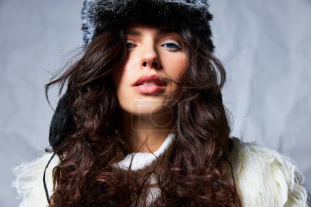 snow queen, curly brunette woman in furry snowy hat and sweater looking at camera on grey backdrop