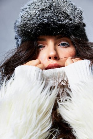 stunning woman in white faux fur jacket, hat and sweater posing on grey backdrop, winter fashion