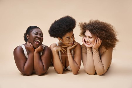 smiling multiethnic body positive women in lingerie lying down and smiling at each other on beige