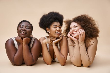 smiling multiethnic body positive women in lingerie lying down and posing on beige, natural beauty