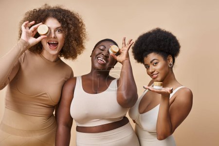 glückliche multiethnische Plus-Size-Frauen in Dessous posieren mit Gläschen kosmetischer Creme auf beige, Hautpflege