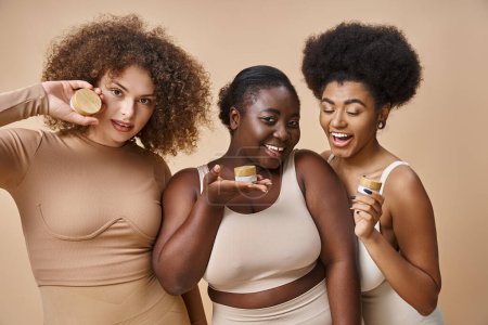 glückliche multiethnische Plus-Size-Frauen in Dessous posieren mit Gläschen kosmetischer Creme auf beige, Hautpflege