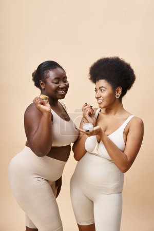 happy african american plus size women in lingerie holding cosmetic cream on beige, skincare