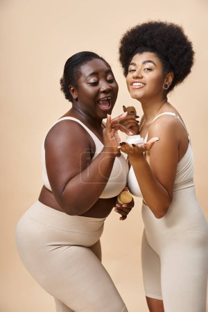 lebenslustige afrikanisch-amerikanische Plus-Size-Frauen in Dessous posieren mit kosmetischer Creme auf beige, Hautpflege