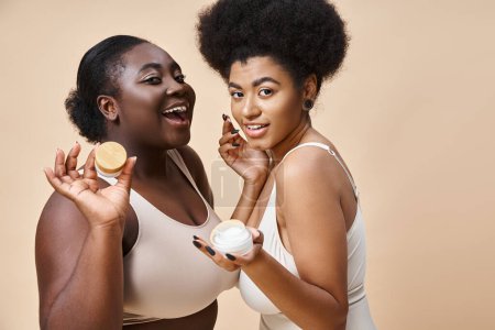 joyful african american plus size women in lingerie posing with cosmetic cream on beige, skincare