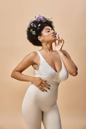 mujer afroamericana sensual con flores de colores en el pelo posando en lencería sobre fondo beige