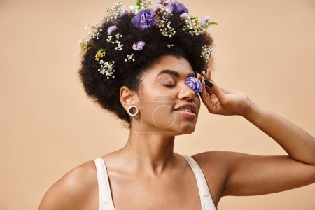 happy african american woman with floral decor in hair and flower near face on beige, natural beauty