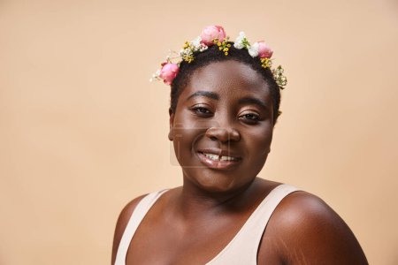 portrait of curvy and happy african american woman with flowers in hair looking at camera on beige