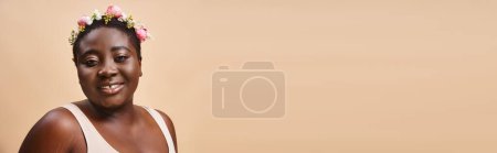curvy and happy african american woman with flowers in hair looking at camera on beige, banner